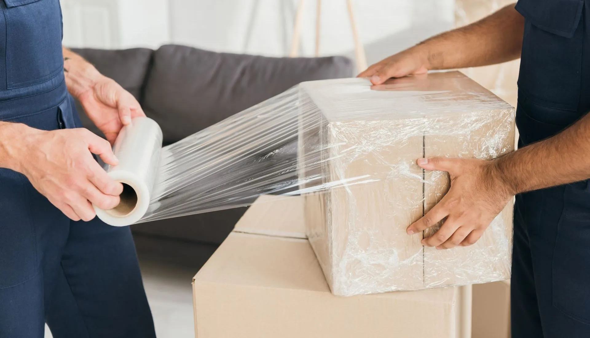 Two people wrapping a cardboard box in clear plastic wrap on moving day.
