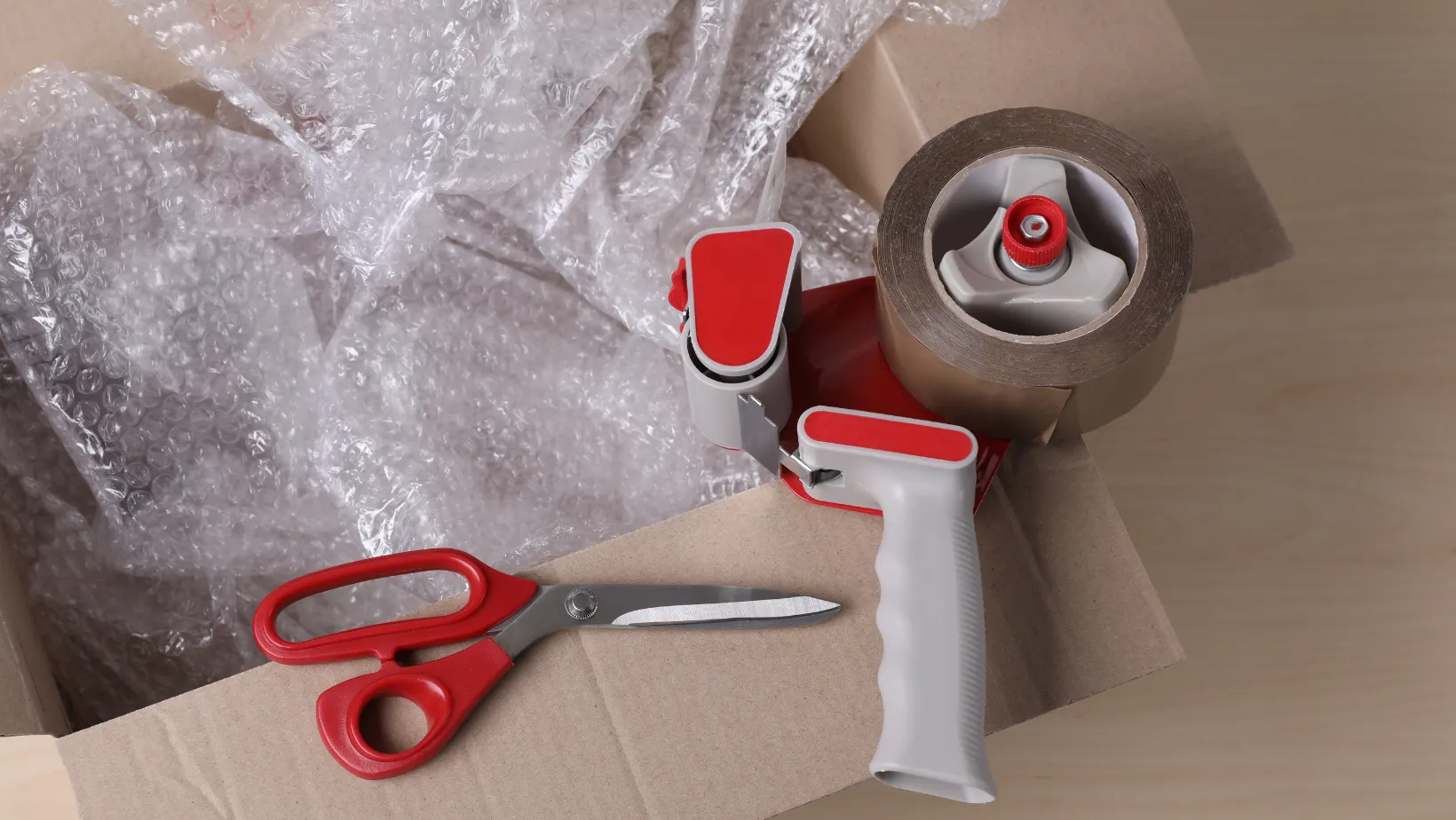 Cardboard box with packing supplies: bubble wrap, tape dispenser, and scissors.